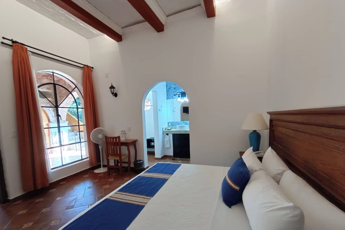 Een slaapkamer met een bed, een houten hoofdbord, een ventilator, een tafel met een stoel en een uitzicht op een badkamer door een boog.