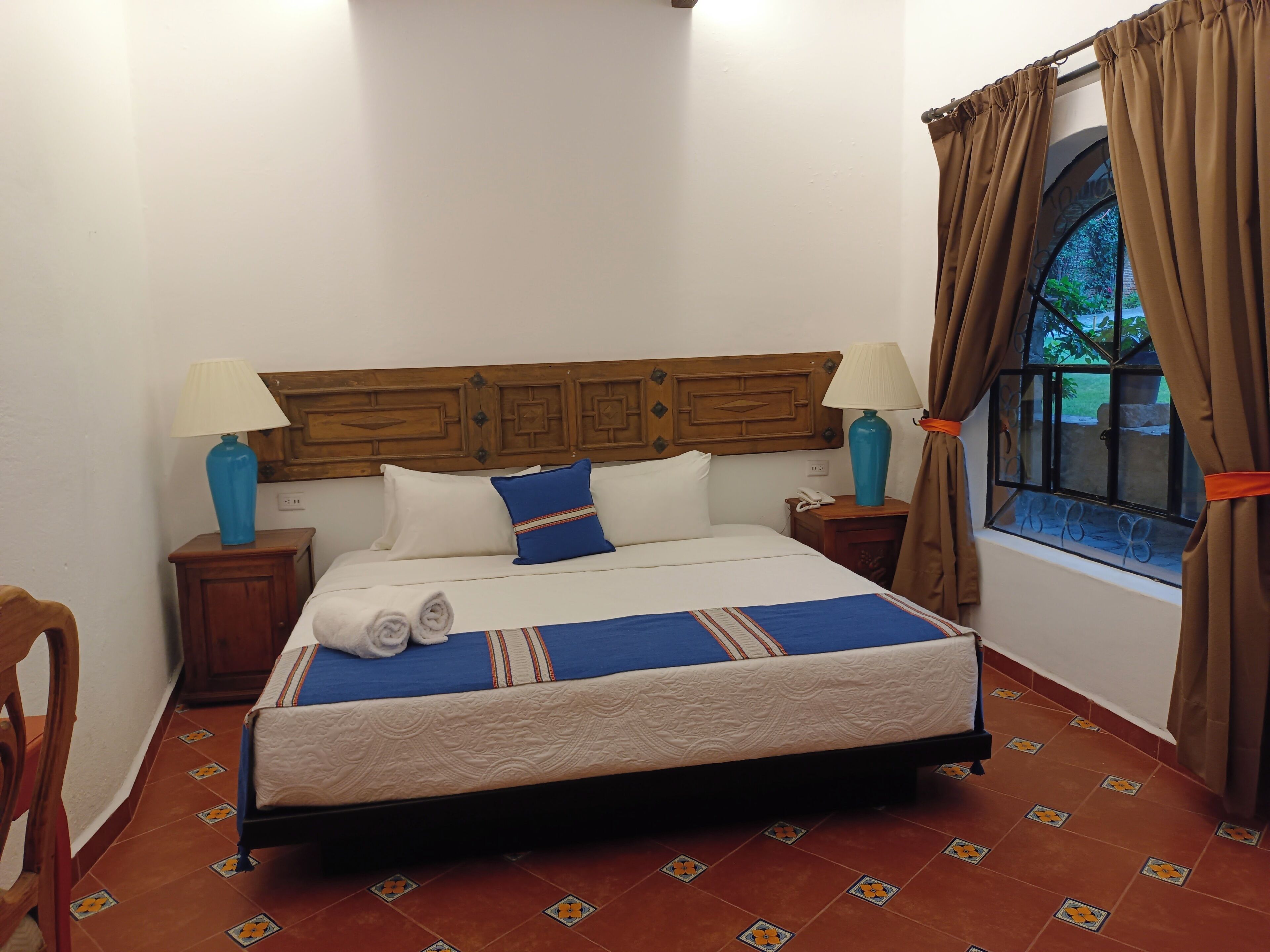 A neatly made bed with a blue and white bedspread, two pillows, and a wooden headboard. There are two bedside tables with lamps, and a window with curtains.