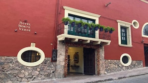 Un bâtiment rouge avec un socle en pierre, un balcon orné de plantes en pot et une enseigne indiquant « Hôtel Monte Verde ».