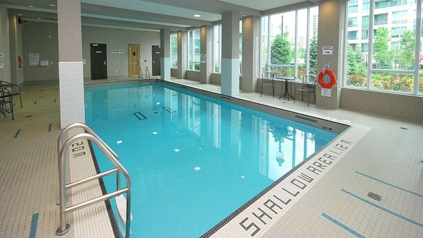 An indoor swimming pool with clear water, a shallow area marked, and a lifebuoy on the wall. - Maplewood Furnished Suites (Mississauga)