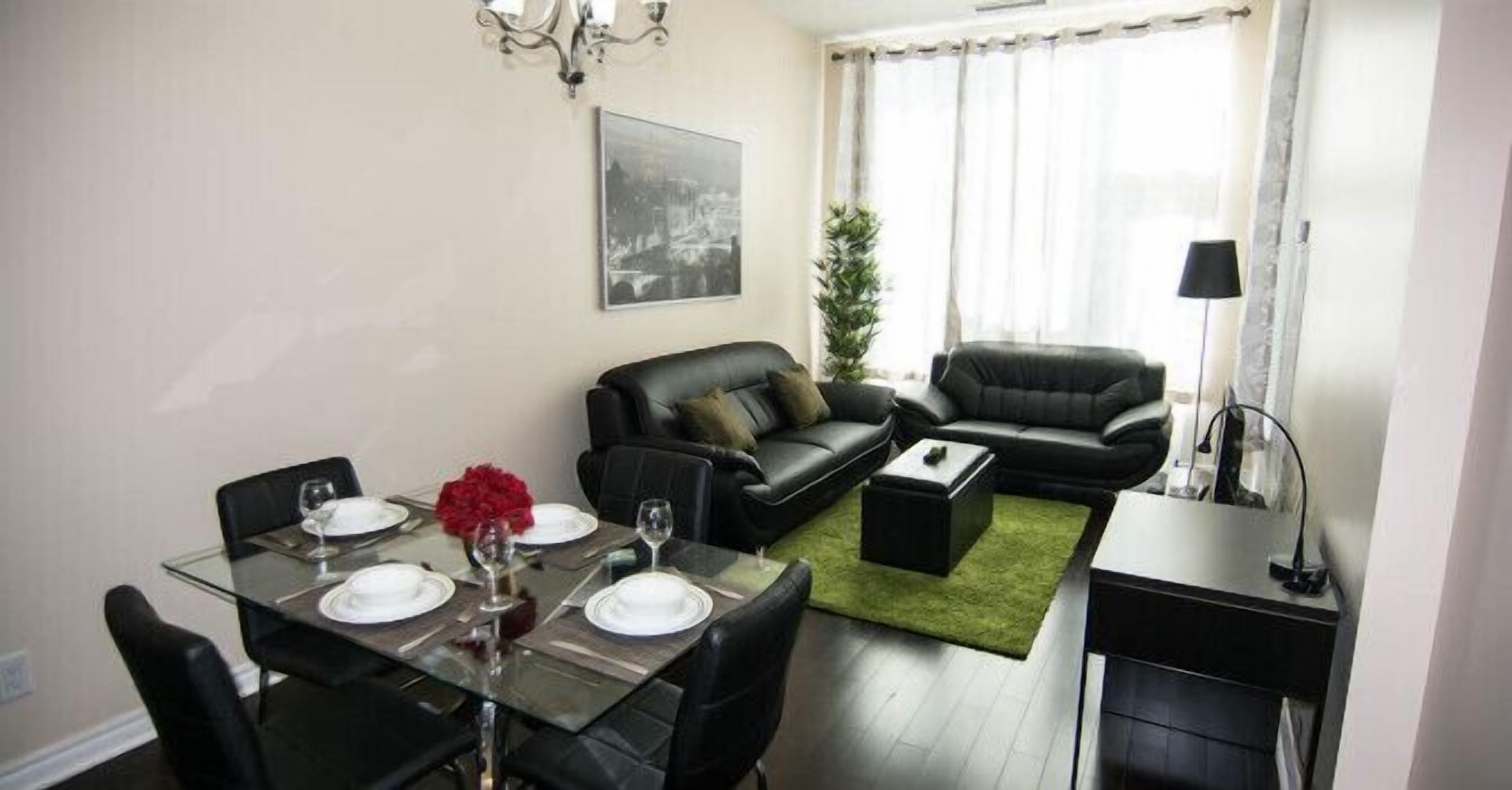A modern living room with a glass dining table set for four, a black leather sofa, and a small side table.