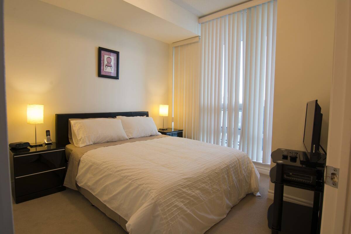A hotel room with a large bed, two bedside tables with lamps, a flat-screen TV, and a window with blinds.