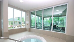 A large indoor swimming pool with a hot tub, surrounded by large windows offering views of outdoor gym equipment and greenery. - Maplewood Furnished Suites (Mississauga)