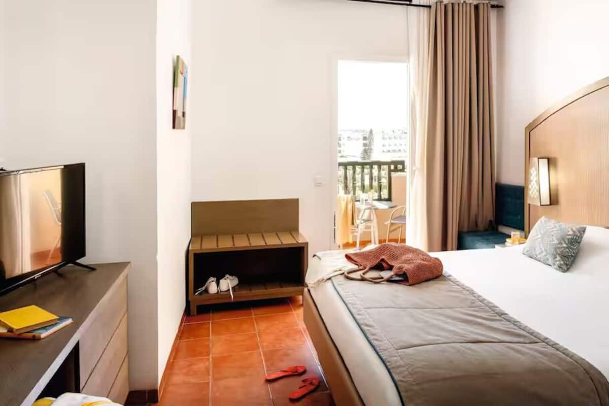 A hotel room with a bed, a desk with a TV, a small seating area, and a balcony with a table and chairs.