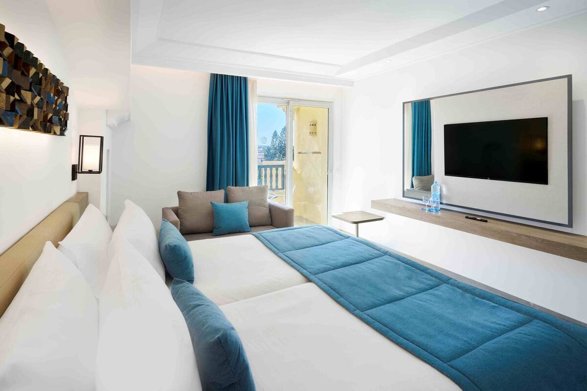 A modern hotel room with a large bed, a flat-screen TV, a balcony with a glass door, and a wall-mounted TV.