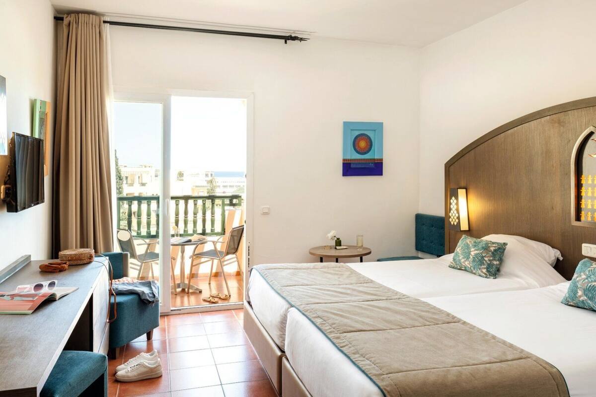 A hotel room with a bed, a desk, a chair, a small table with a vase, and a balcony with a view.