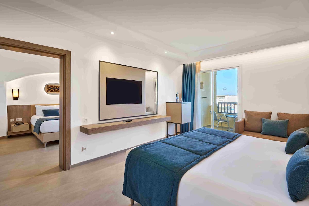 A modern hotel room with a large bed, a flat-screen TV mounted on the wall, a balcony with a view, and a separate seating area with a sofa and armchairs.
