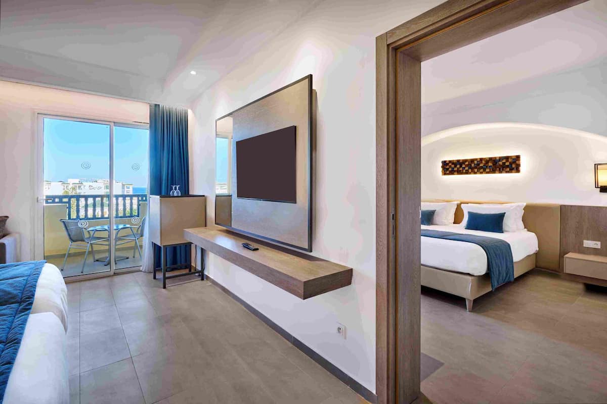 A modern hotel room with a large TV, a balcony with a table and chairs, a bed with a headboard, and a bedside table.