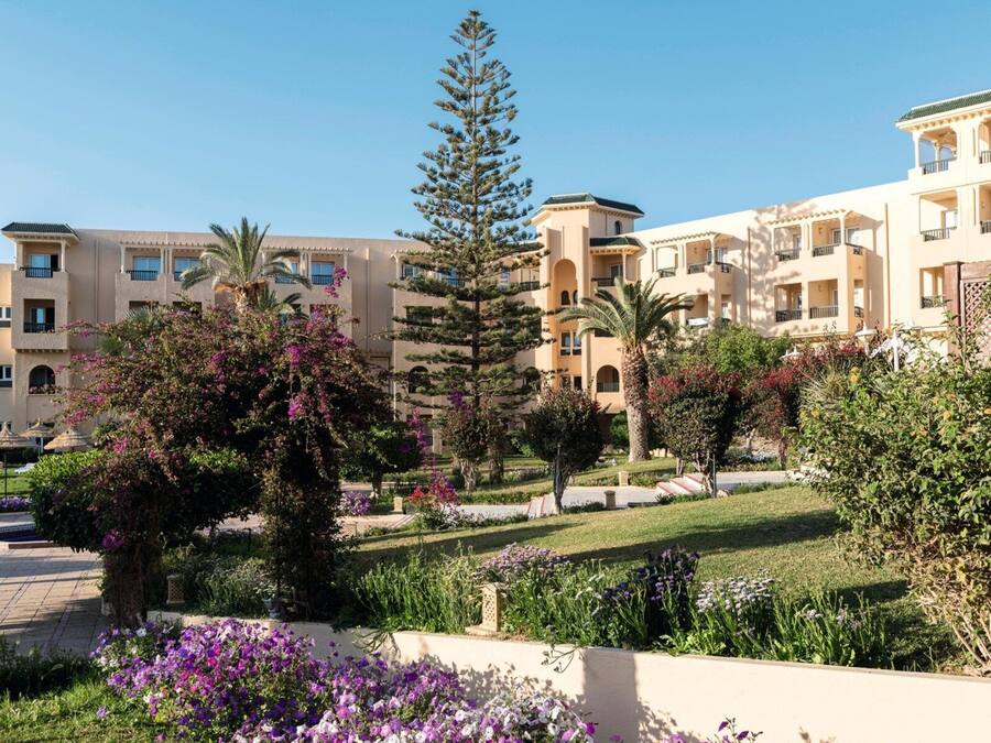 A hotel with multiple balconies, a well-maintained garden with various plants and flowers, and a clear sky.