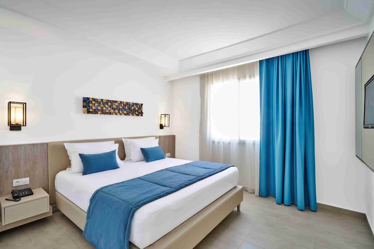 A modern hotel room with a large bed, two bedside tables, a wall-mounted artwork, and a large window with blue curtains.
