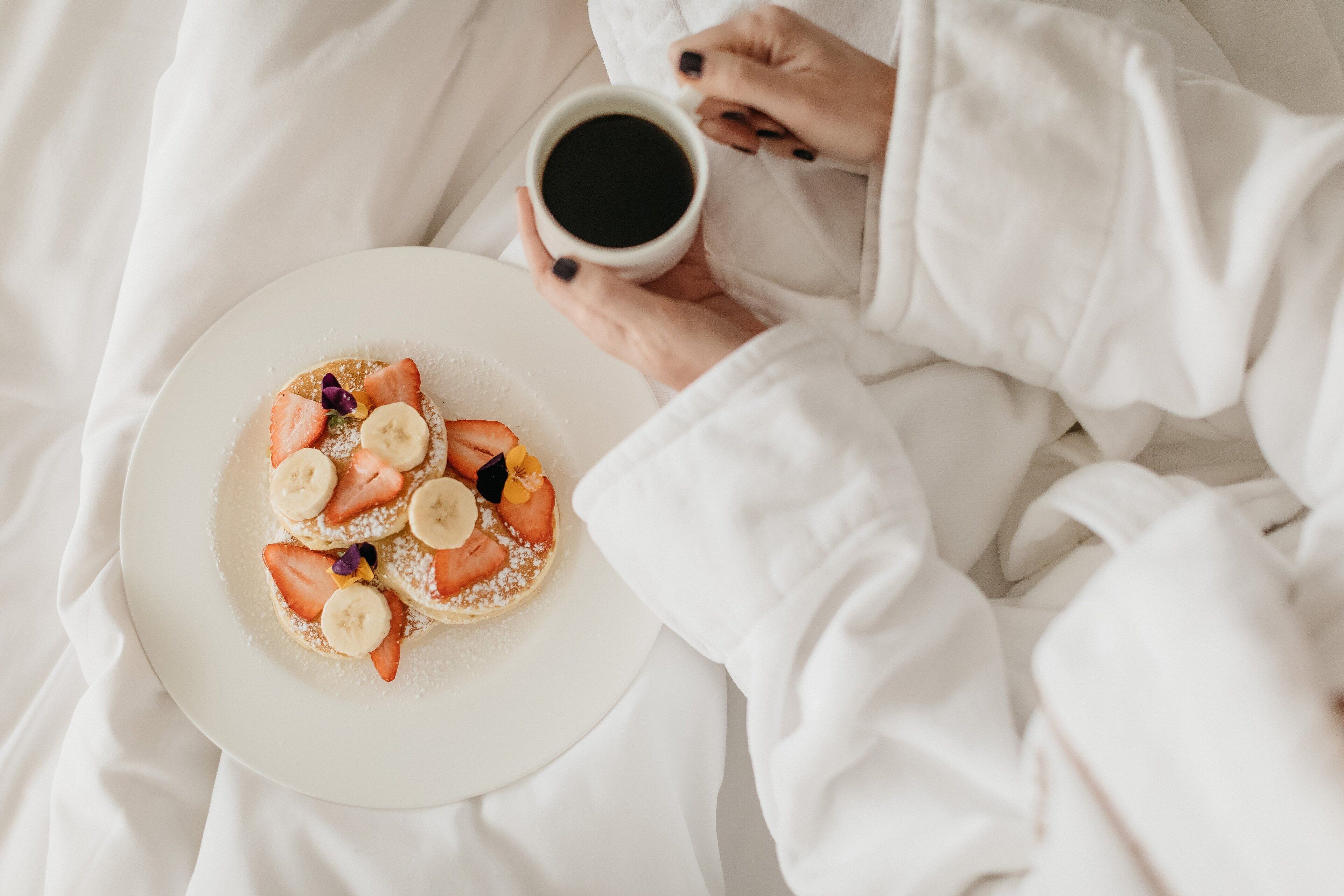 A person in white robes holding a cup of coffee and a plate with pancakes topped with strawberries and bananas.
