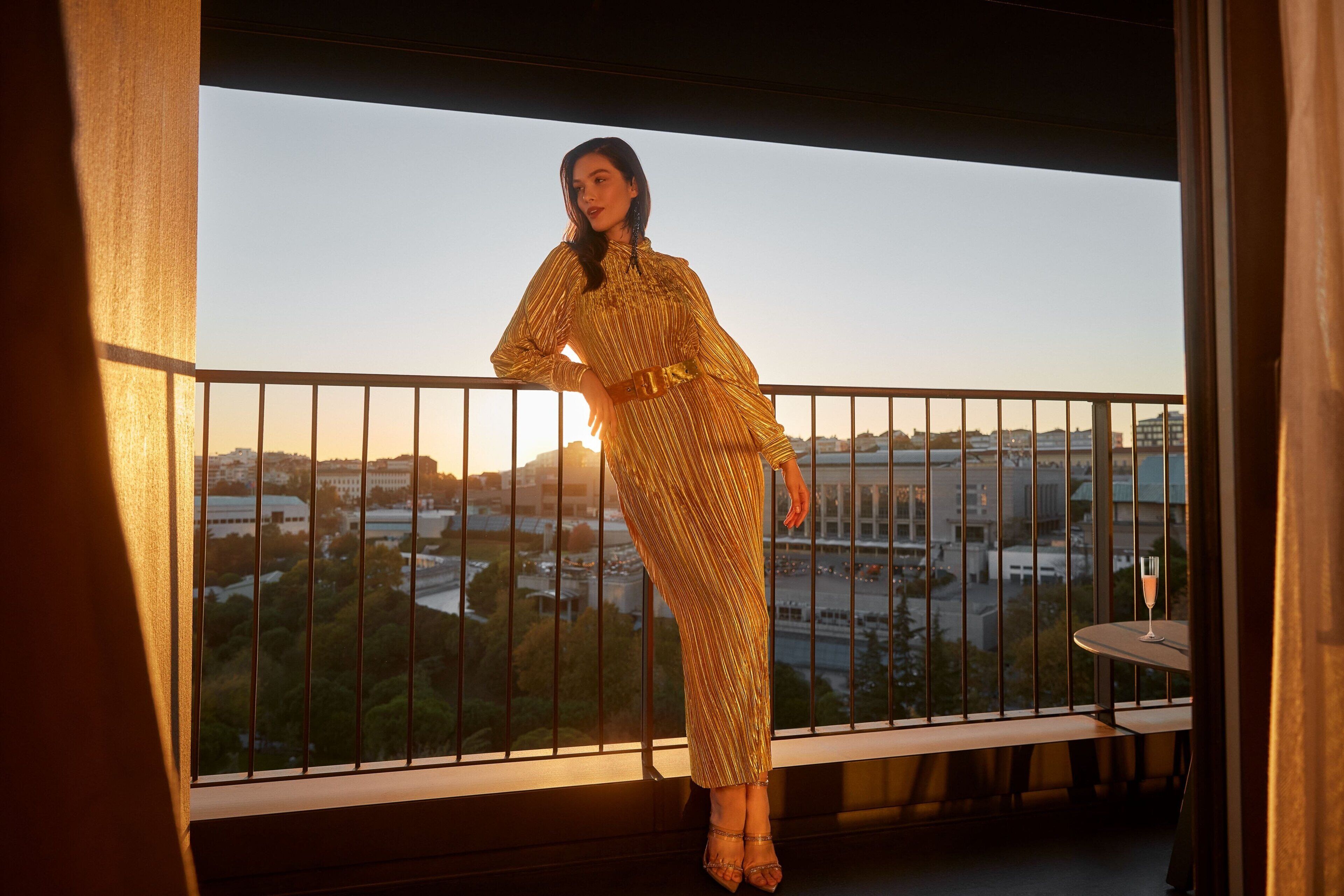 A person in a patterned dress on a balcony with a view of a cityscape and a table with a glass of drink.