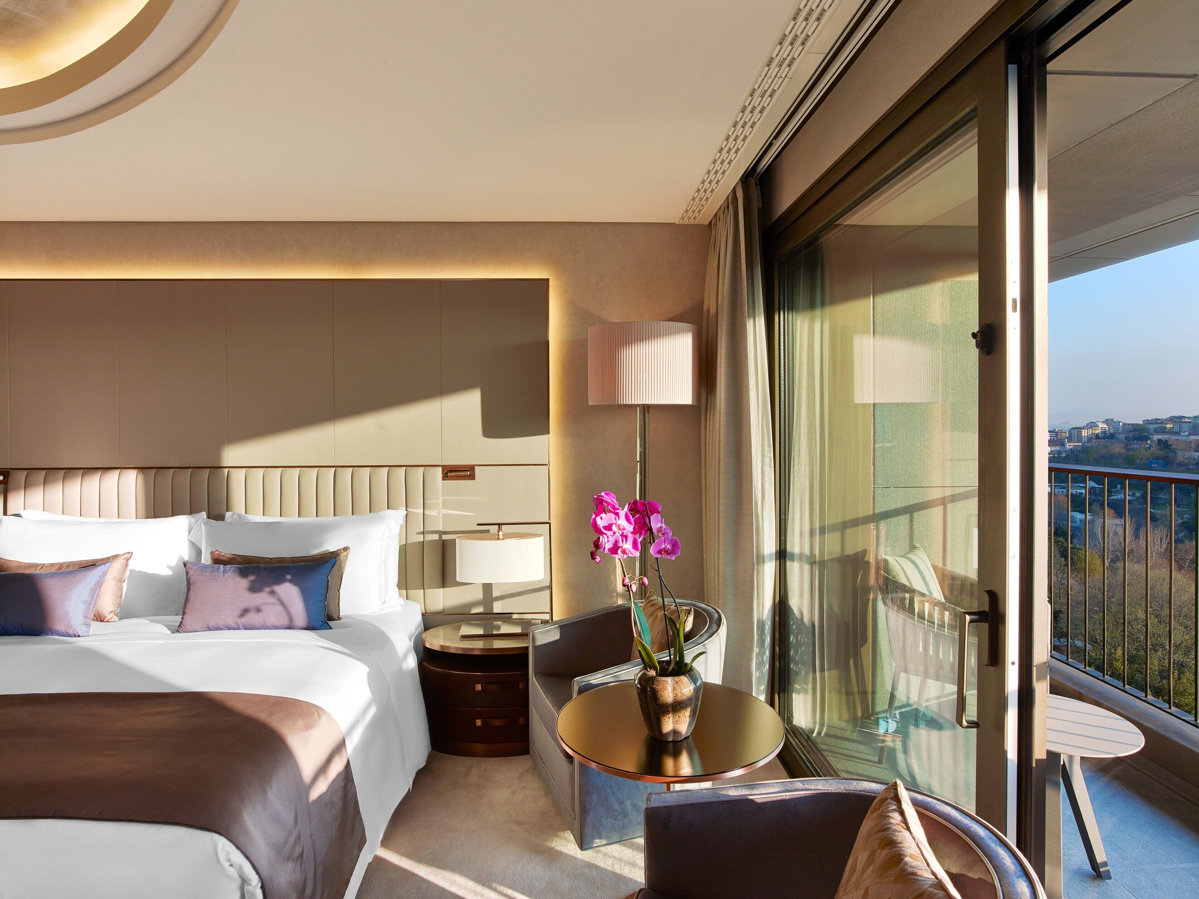 Een hotelkamer met een bed, nachtkastje, lamp, stoel en een balkon met uitzicht.