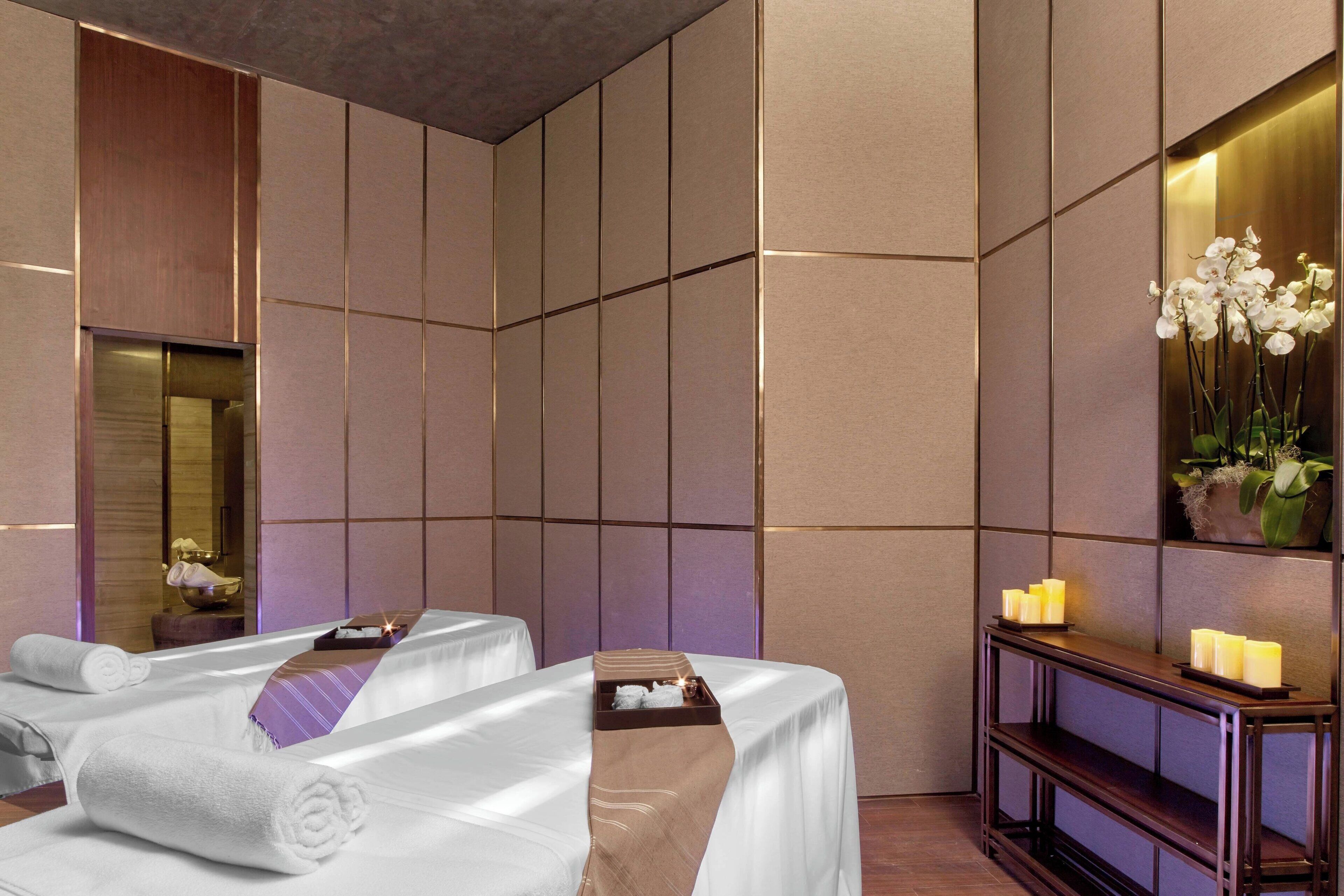 A spa room with two massage tables, a wooden console table with candles, and a large mirror on the wall.