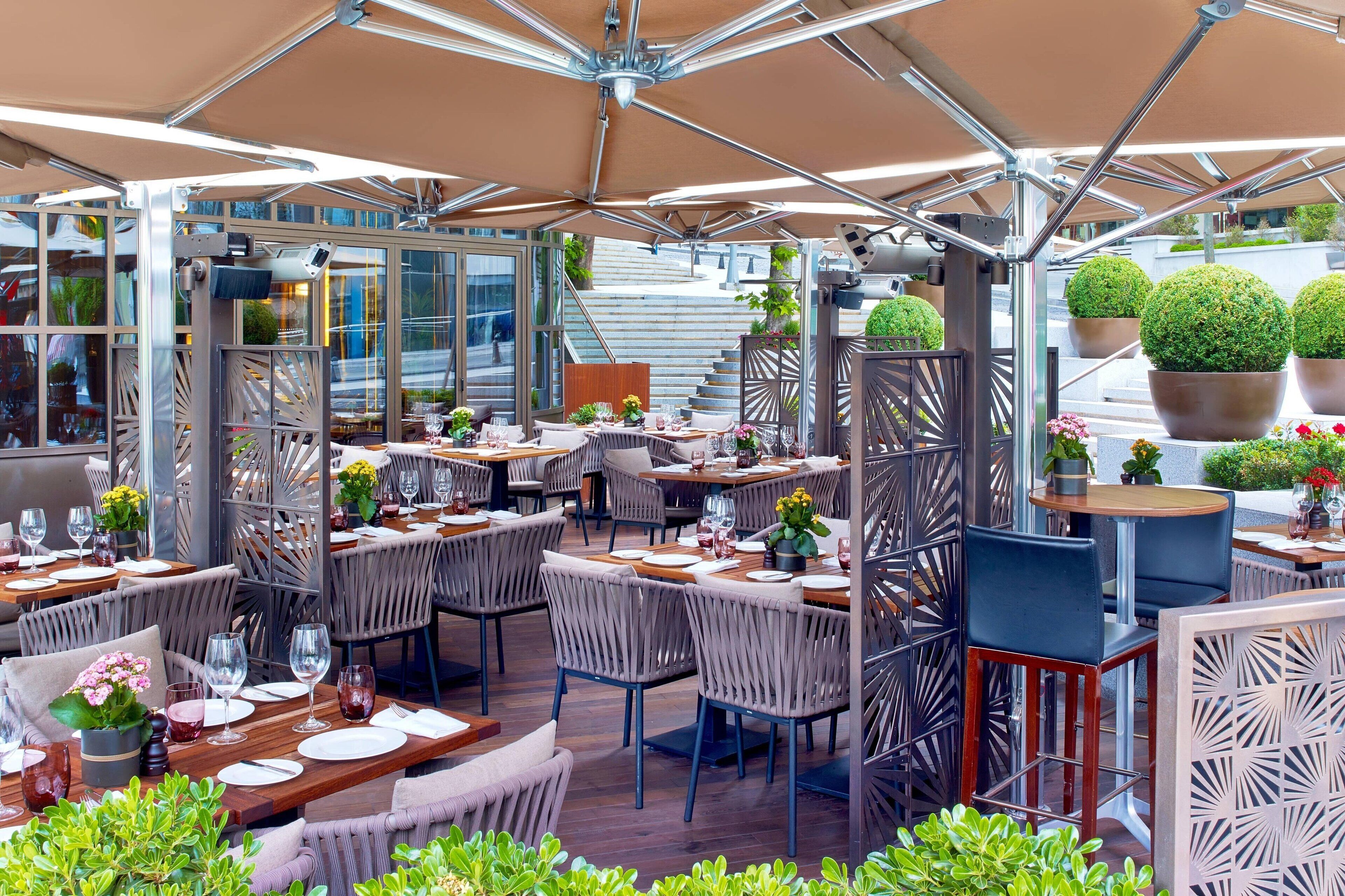 An outdoor dining area with multiple tables and chairs, surrounded by glass partitions and potted plants.