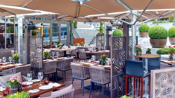 An outdoor dining area with multiple tables and chairs, surrounded by glass partitions and potted plants.