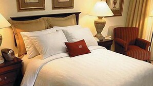 Una cama bien hecha con sábanas blancas y una almohada roja decorativa, una mesita de noche de madera con una lámpara y un sillón a rayas.