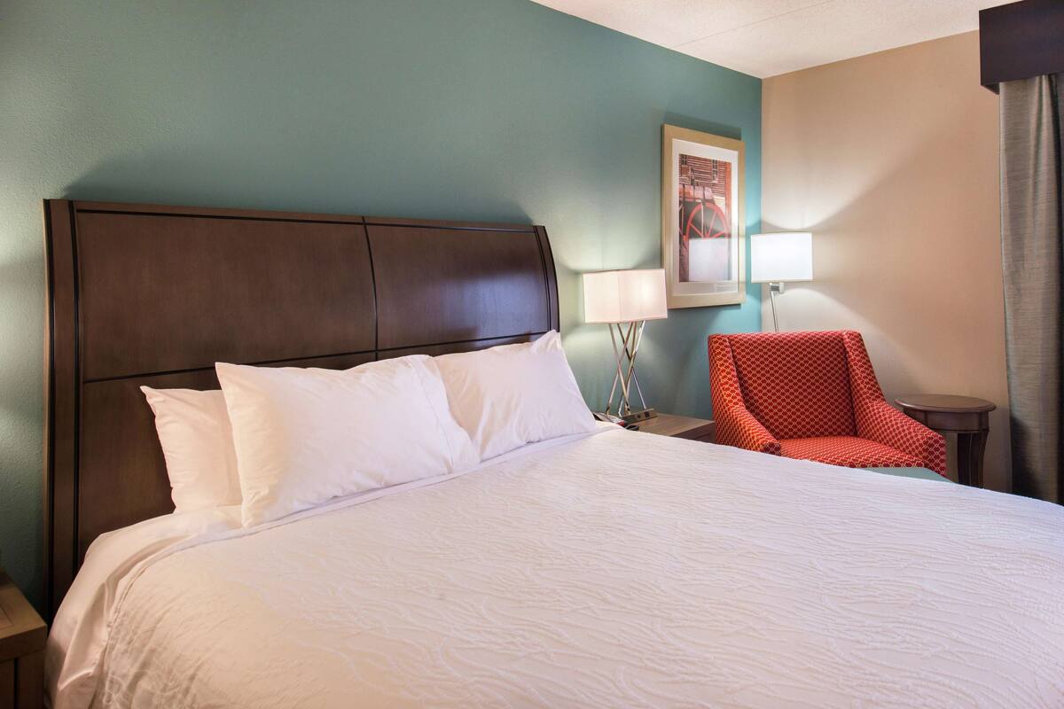 A hotel room with a large bed, a wooden headboard, a red armchair, a small table with a lamp, and a framed picture on the wall.