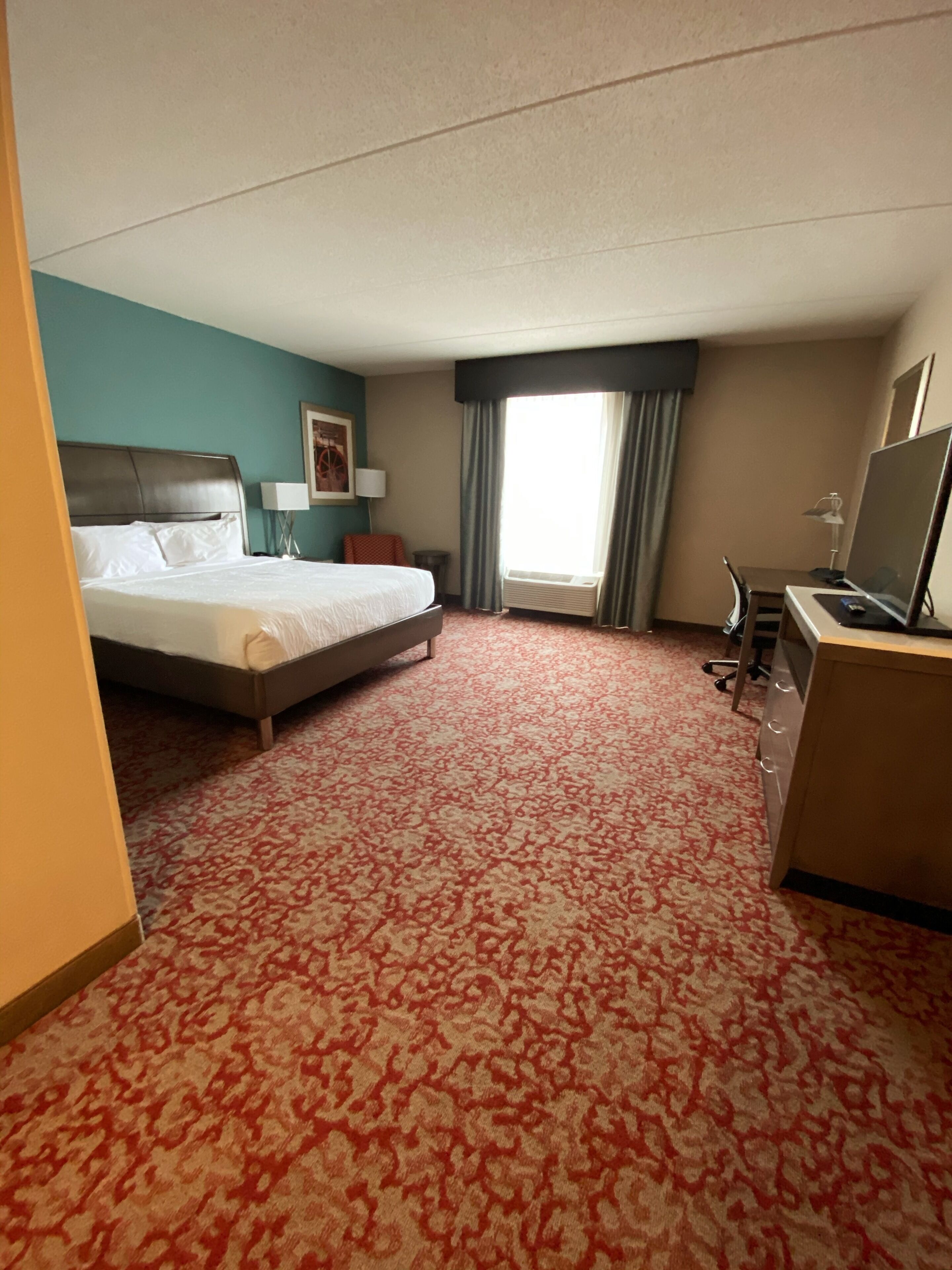 Habitación de hotel con cama, escritorio y un amplio ventanal con cortinas.