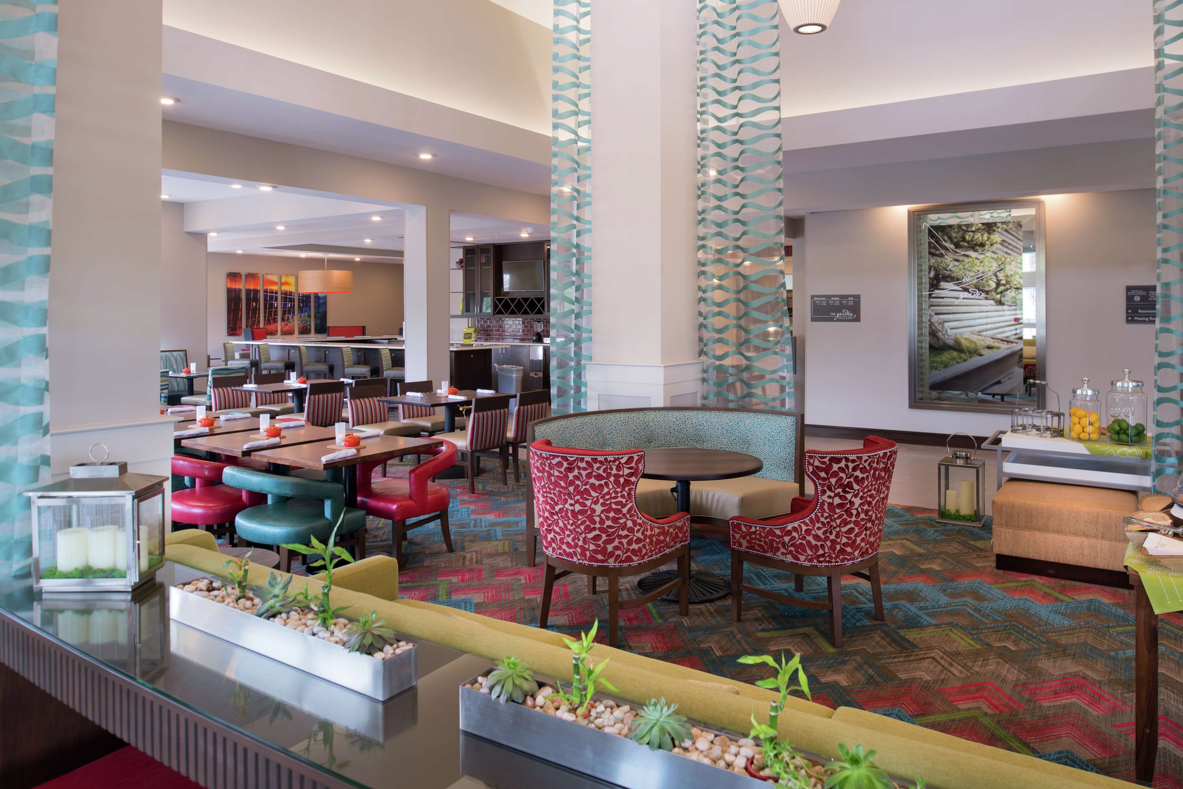 A modern hotel lobby with a central seating area, a bar, and a kitchen in the background.