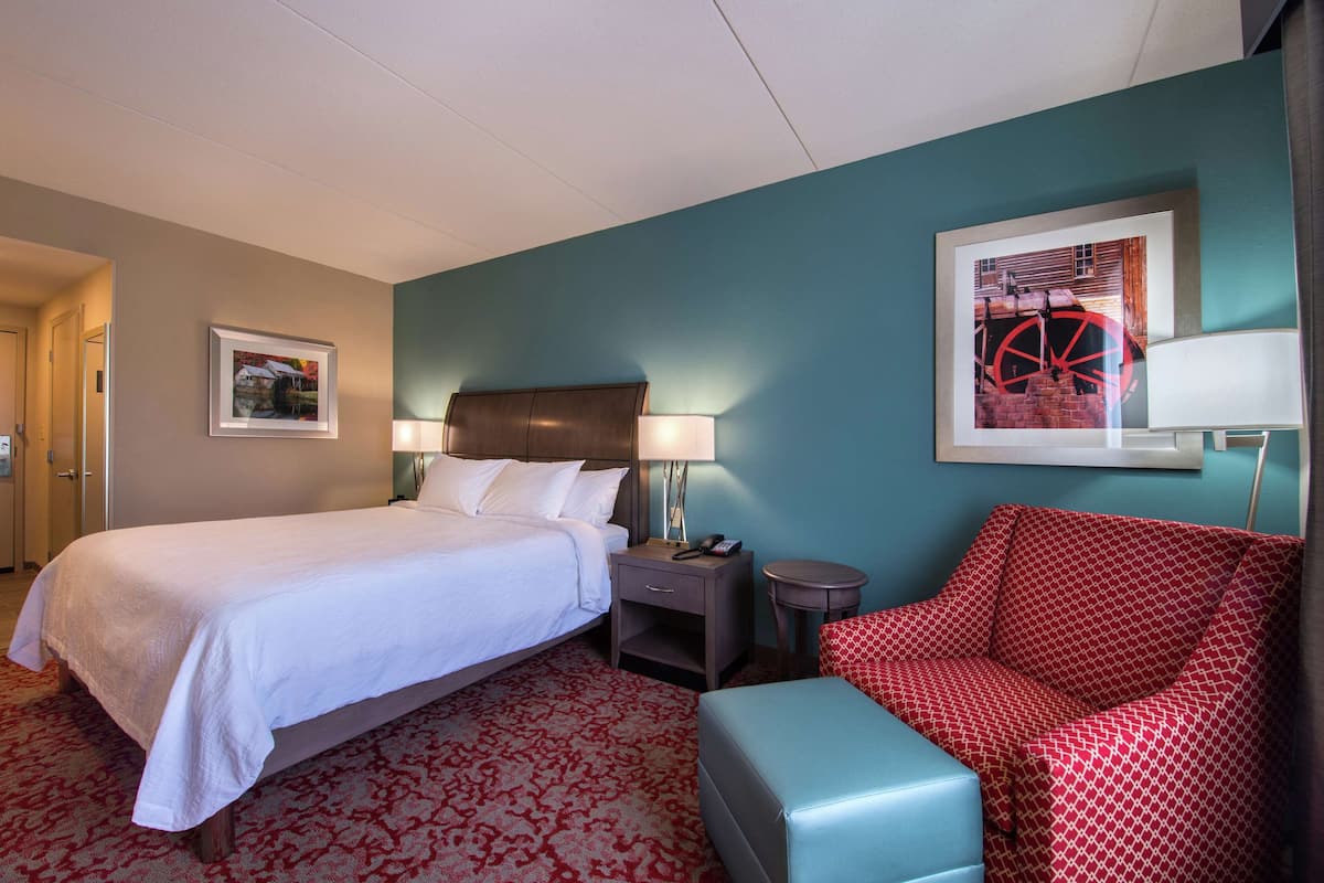 A hotel room with a bed, bedside table, lamp, and a red patterned chair.