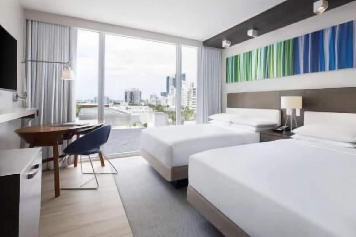 Habitación de hotel con dos camas, un escritorio, una silla y un ventanal con vistas a la ciudad.