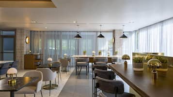 A modern dining area with a long counter, multiple tables, and chairs. There are pendant lights above the counter and a large window with curtains.