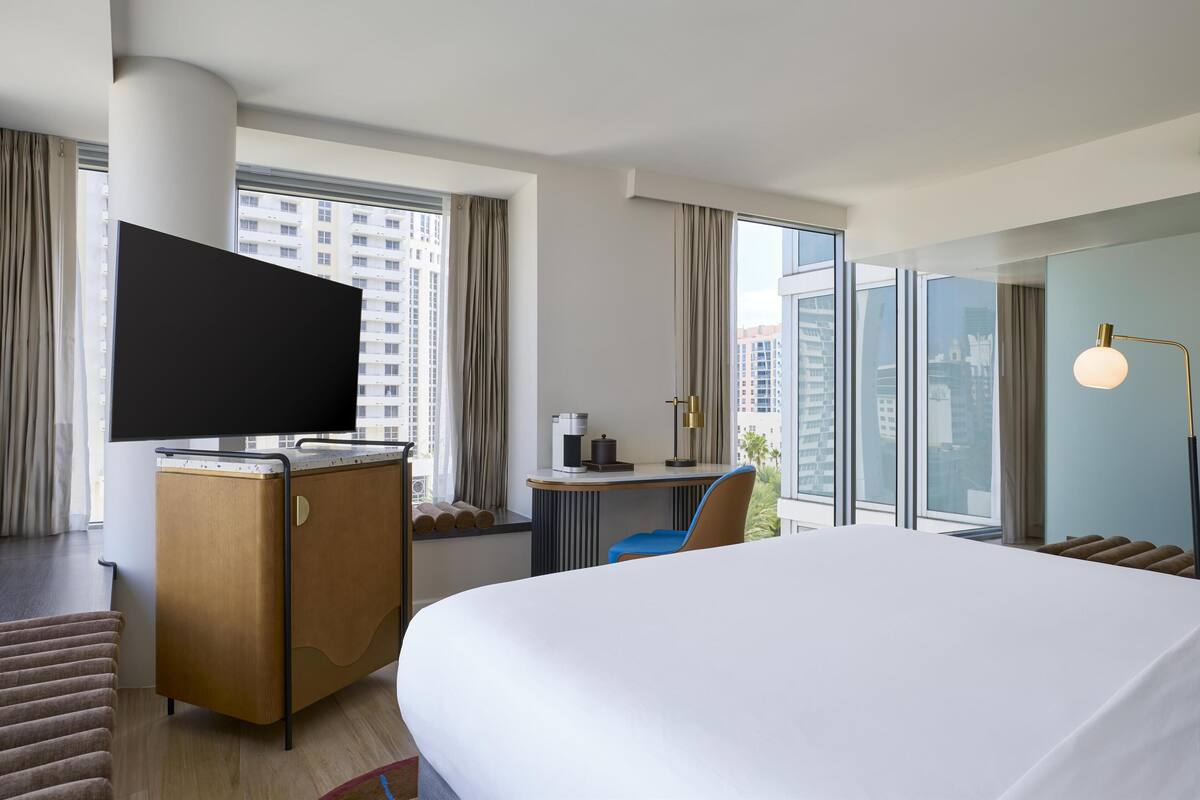 Habitación de hotel con una cama grande, un escritorio con una silla, un televisor de pantalla plana y vistas a la ciudad a través de amplias ventanas.