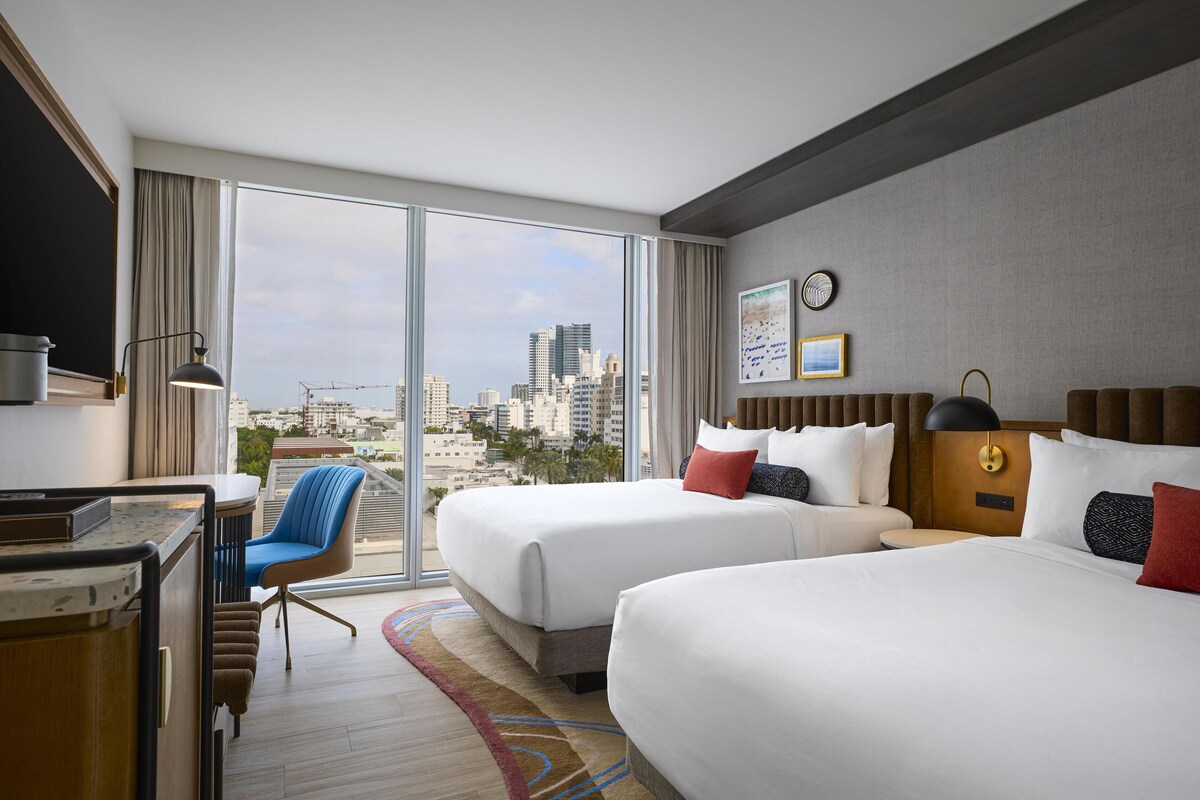 A hotel room with two beds, a desk, a chair, and a large window with a city view.
