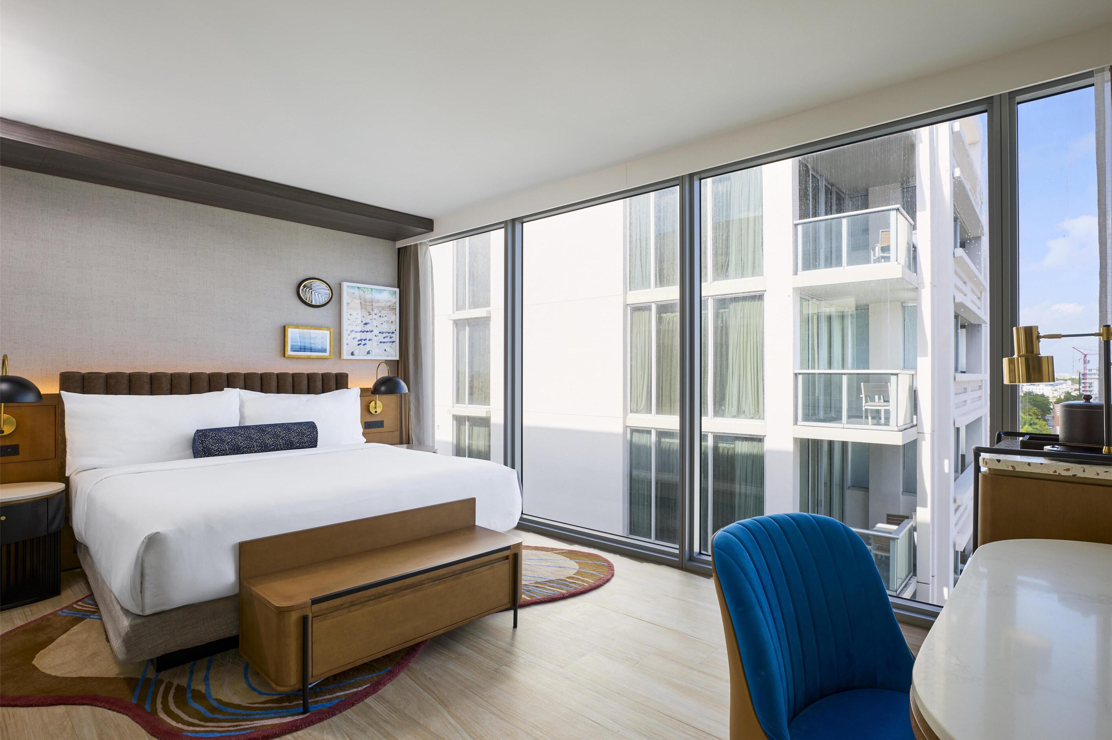 Habitación de hotel moderna con una cama grande, un escritorio y vistas a la ciudad a través de ventanales del suelo al techo.
