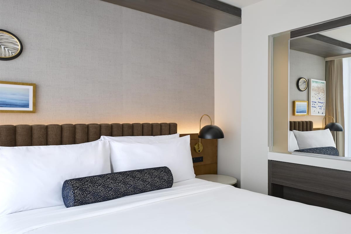 Habitación de hotel ordenada con cama, mesita de noche, lámpara, espejo y reloj en la pared.