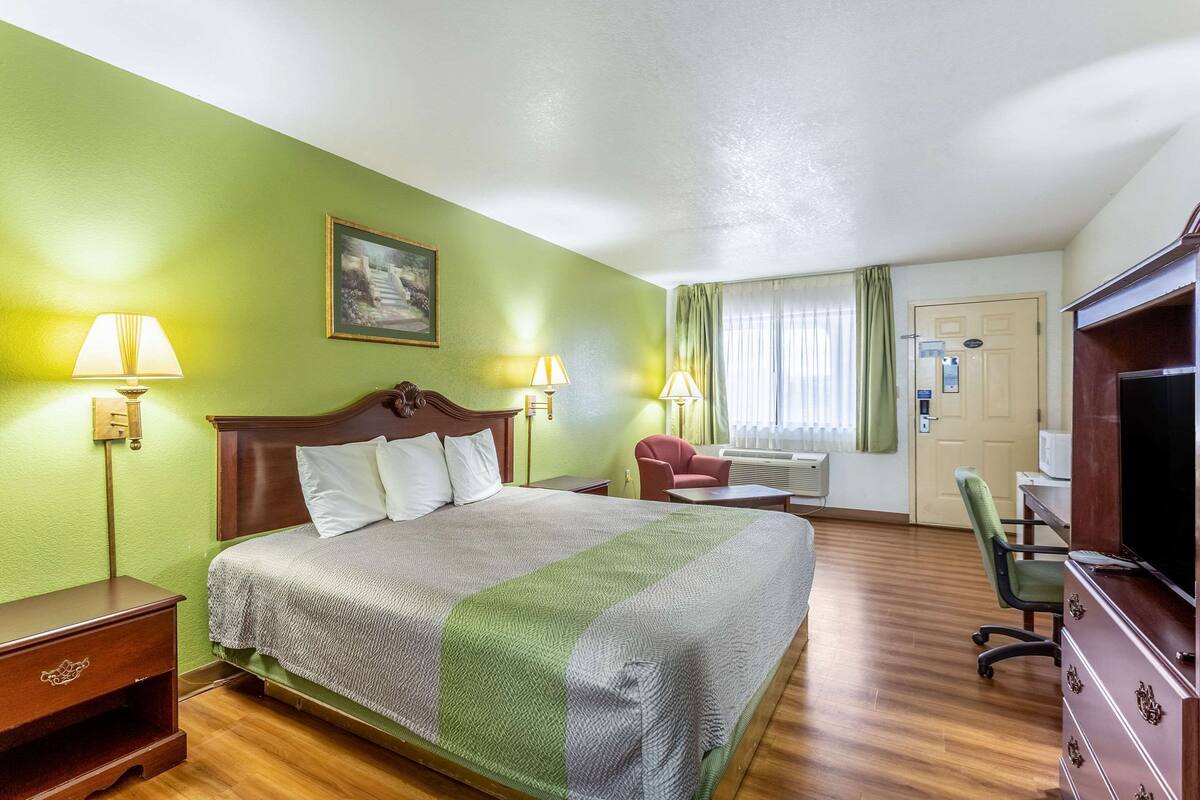 A hotel room with a bed, two bedside lamps, a chair, a desk with a television, and a door with an elevator.