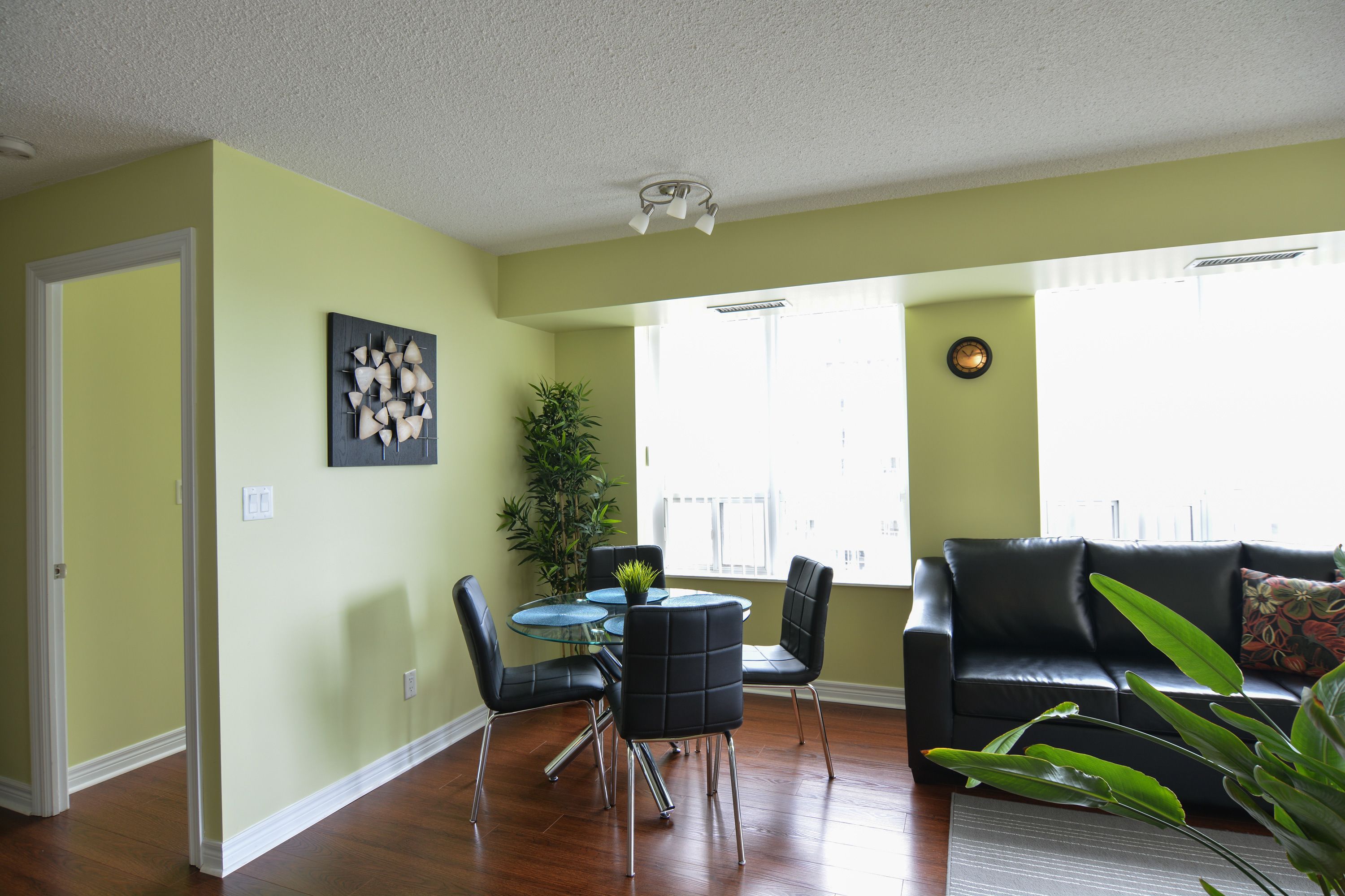 A living room with a black leather sofa, a glass coffee table, and a dining table with chairs. There is a potted plant and a painting on the wall.