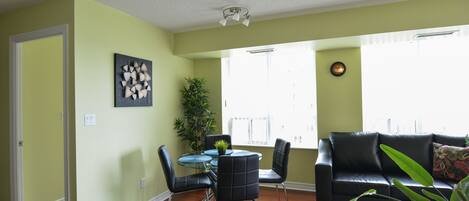 A living room with a black leather sofa, a glass coffee table, and a dining table with chairs. There is a potted plant and a painting on the wall.
