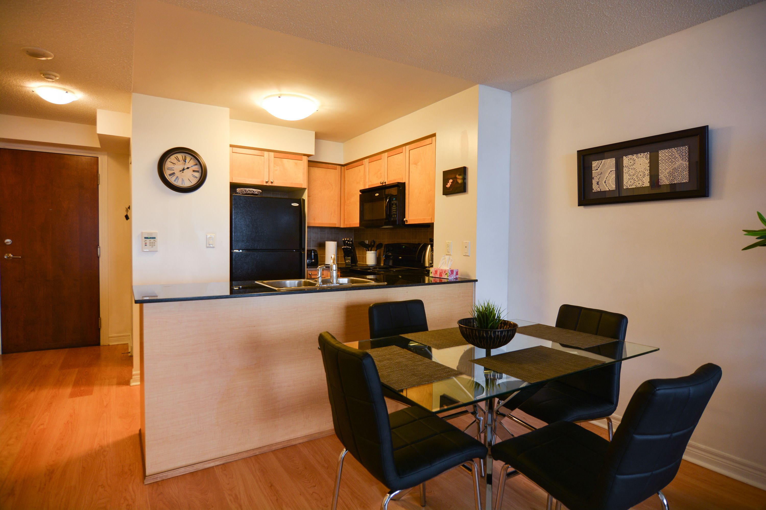 A modern kitchen with wooden cabinets, a black refrigerator, and a microwave. There is a dining area with a glass table and black chairs.