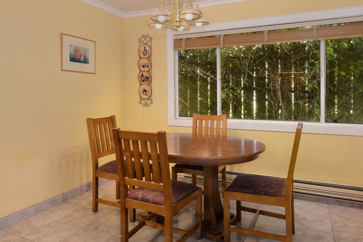A dining area with a round wooden table and four chairs, a framed picture on the wall, and a window with a view of trees.