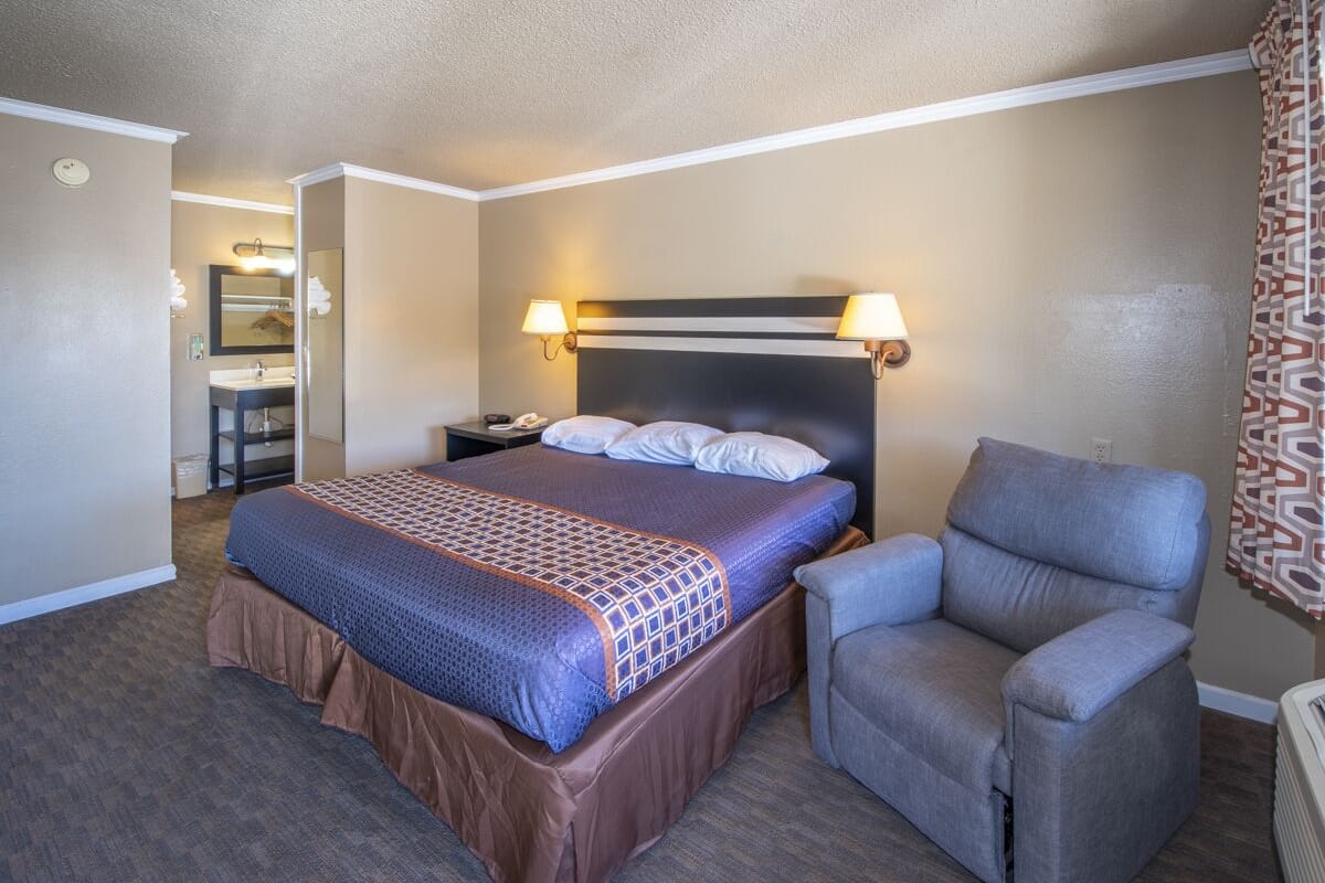 A hotel room with a bed, a chair, a nightstand, and a bathroom visible in the background.