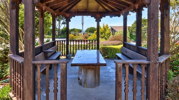 Um gazebo de madeira com um banco e uma mesa, cercado por vegetação.