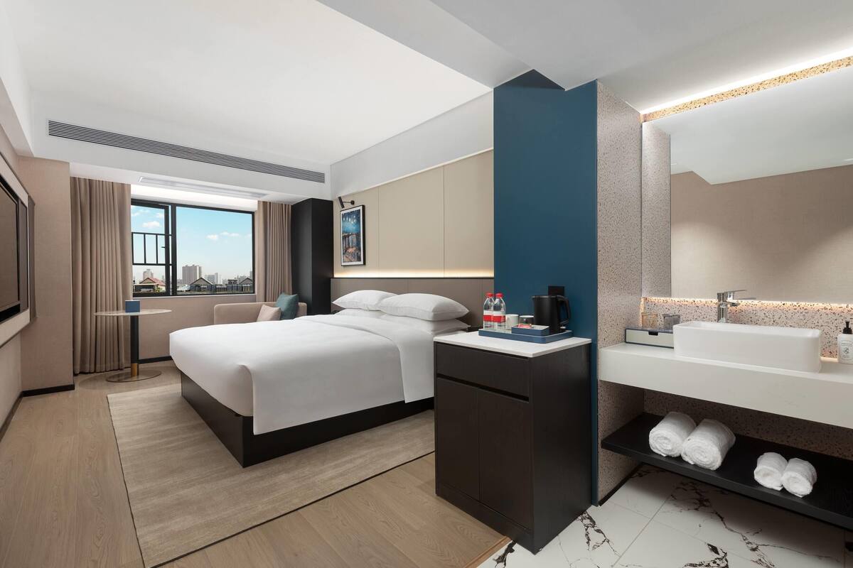 A modern hotel room with a large bed, a desk, a bathroom with a sink and mirror, and a view of the city.
