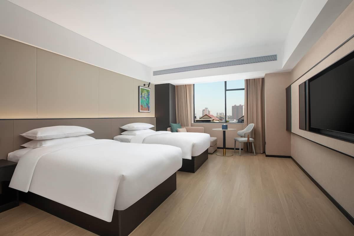 A hotel room with two beds, a desk, a chair, a TV, and a window with a city view.