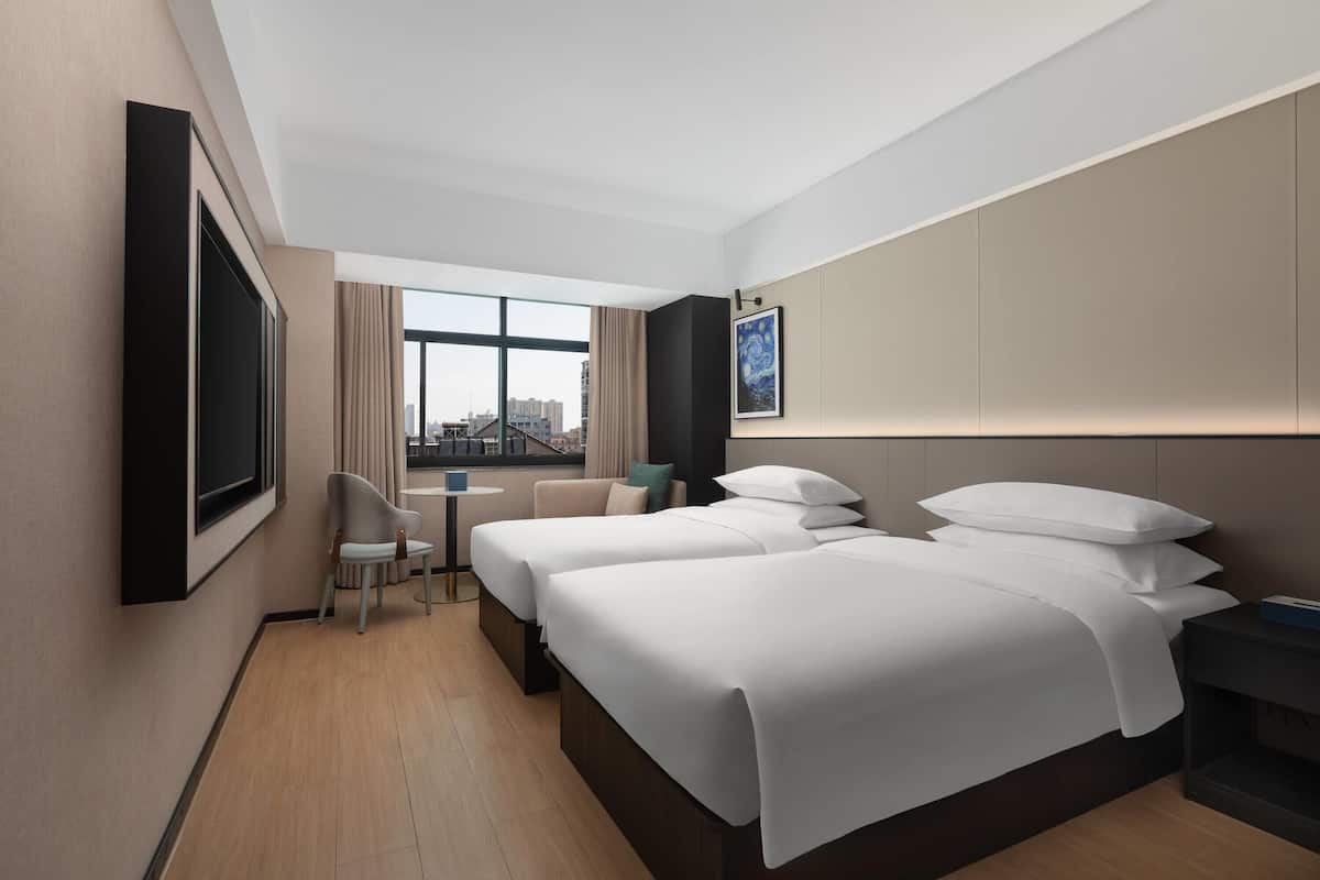 A hotel room with two beds, a TV, a small table, a chair, and a large window with a city view.