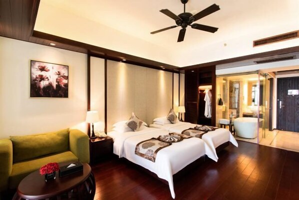 Une chambre d’hôtel avec un lit, un fauteuil vert, une petite table avec un vase et un ventilateur de plafond.