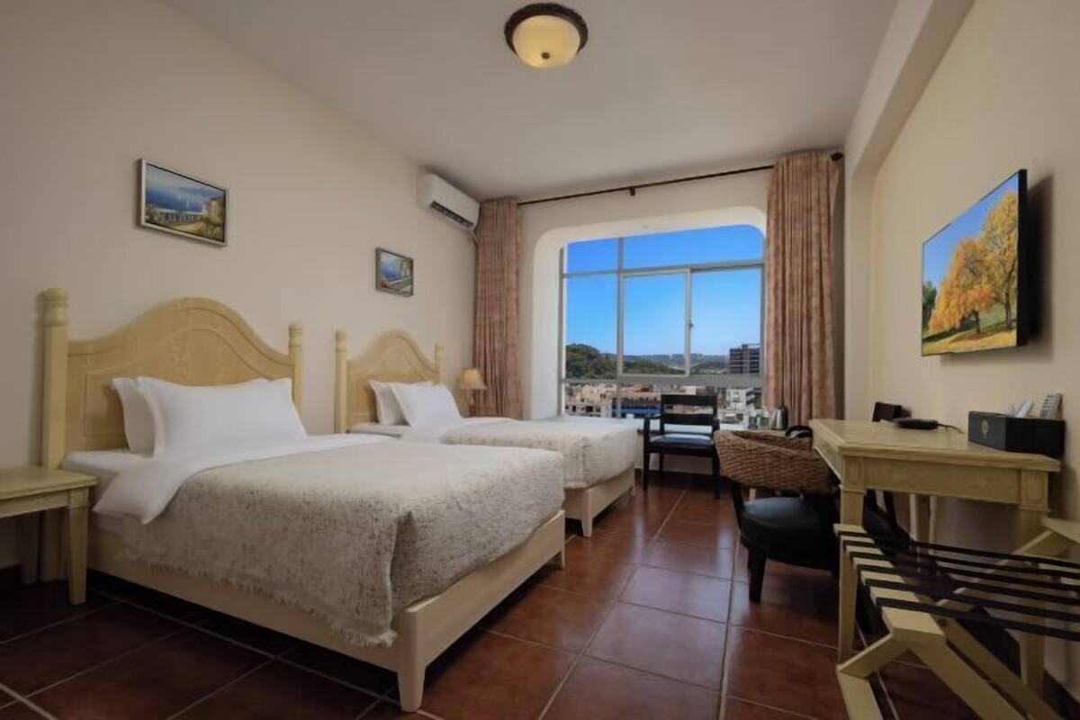 A hotel room with two beds, a desk, and a chair. There is a large window with a view of buildings.
