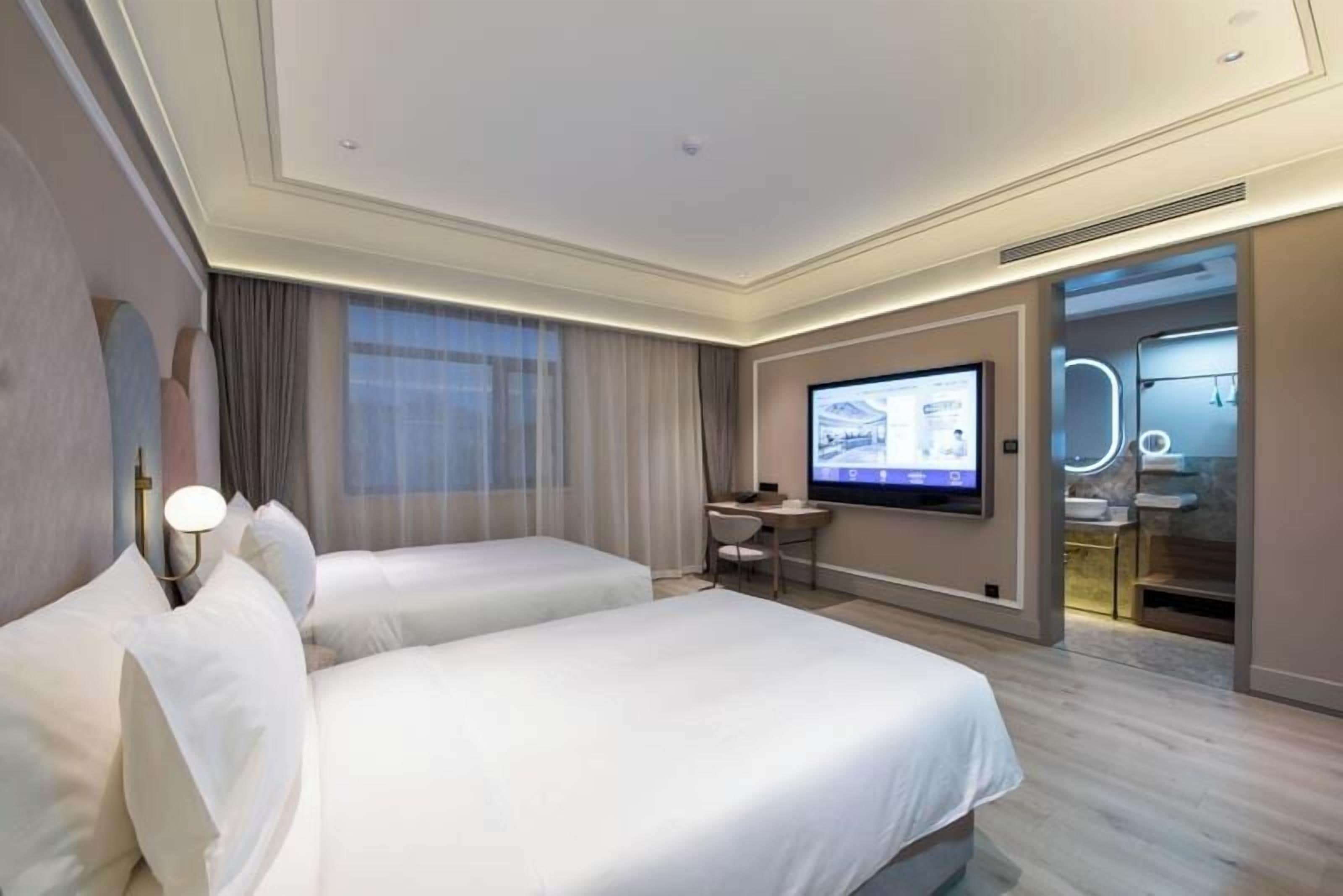 A hotel room with two beds, a desk, a TV, and a bathroom with a mirror and lights.