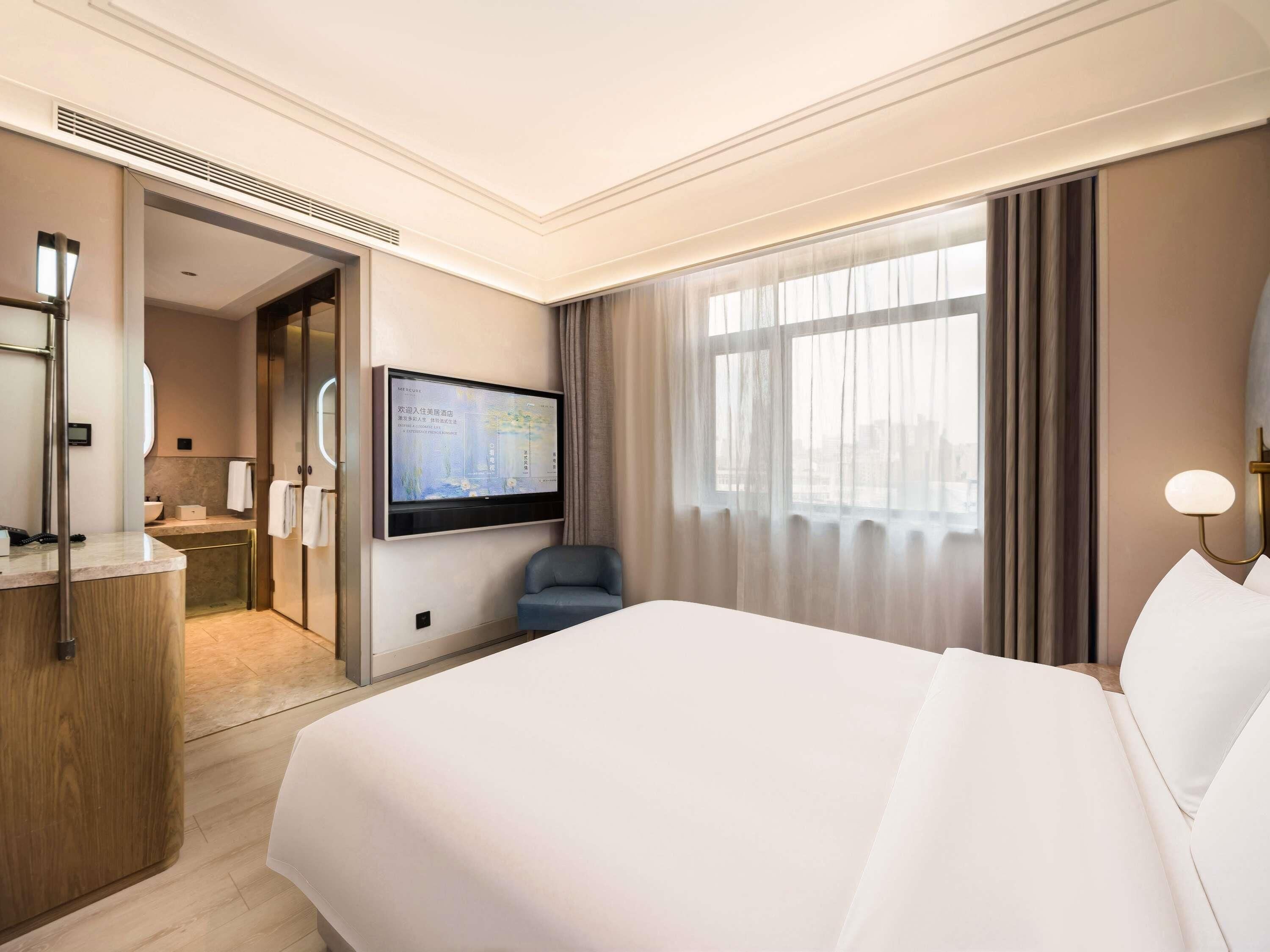 A hotel room with a large bed, a desk with a computer, a TV mounted on the wall, and a window with curtains.