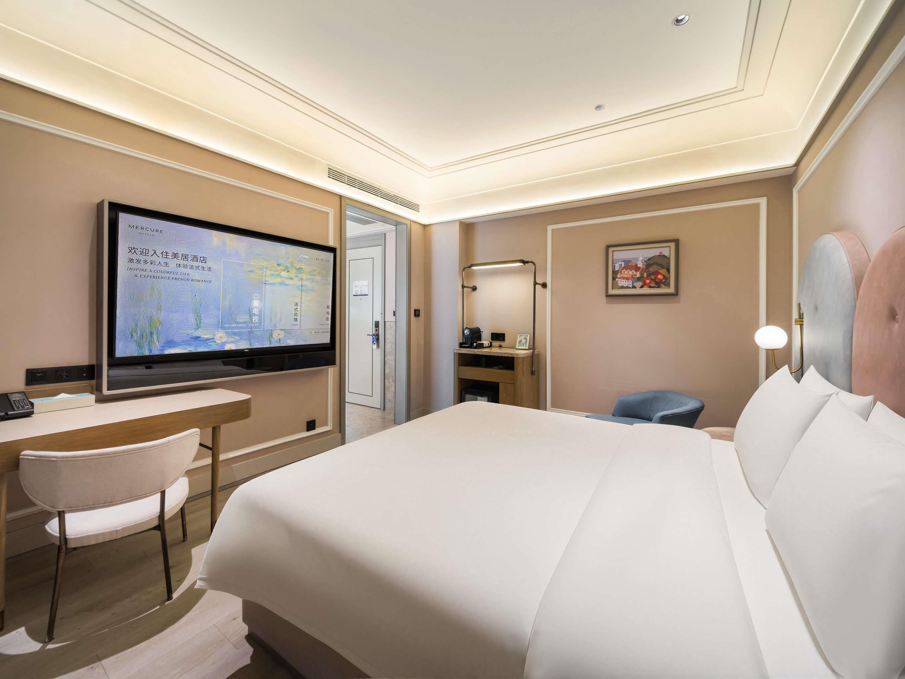 Ein Hotelzimmer mit Bett, Schreibtisch, Flachbildfernseher, Stuhl, Nachttisch und Wandlampe.