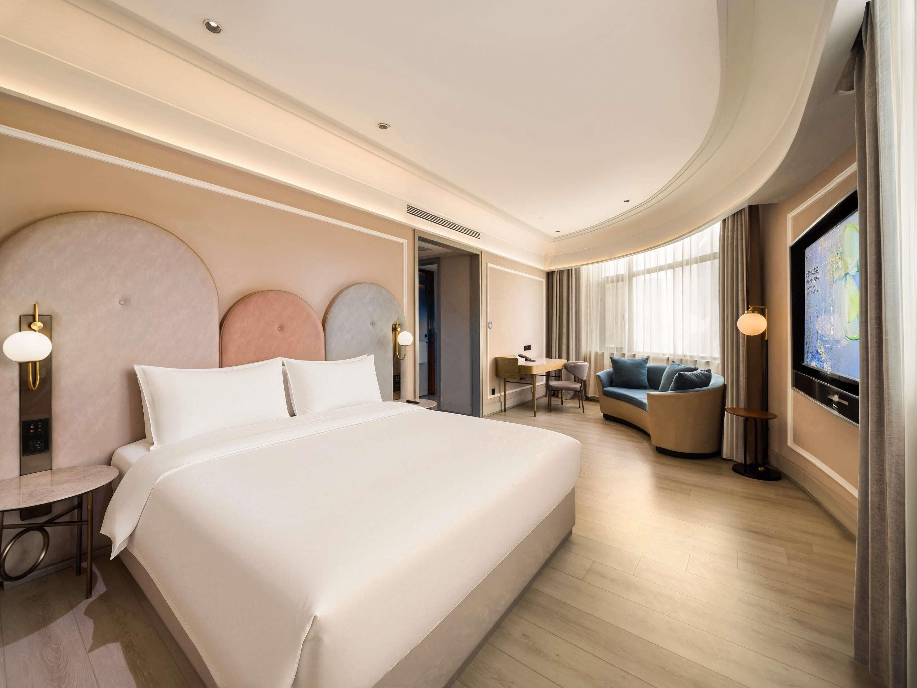 A modern hotel room with a large bed, a seating area with a sofa and armchairs, a desk, and a television.