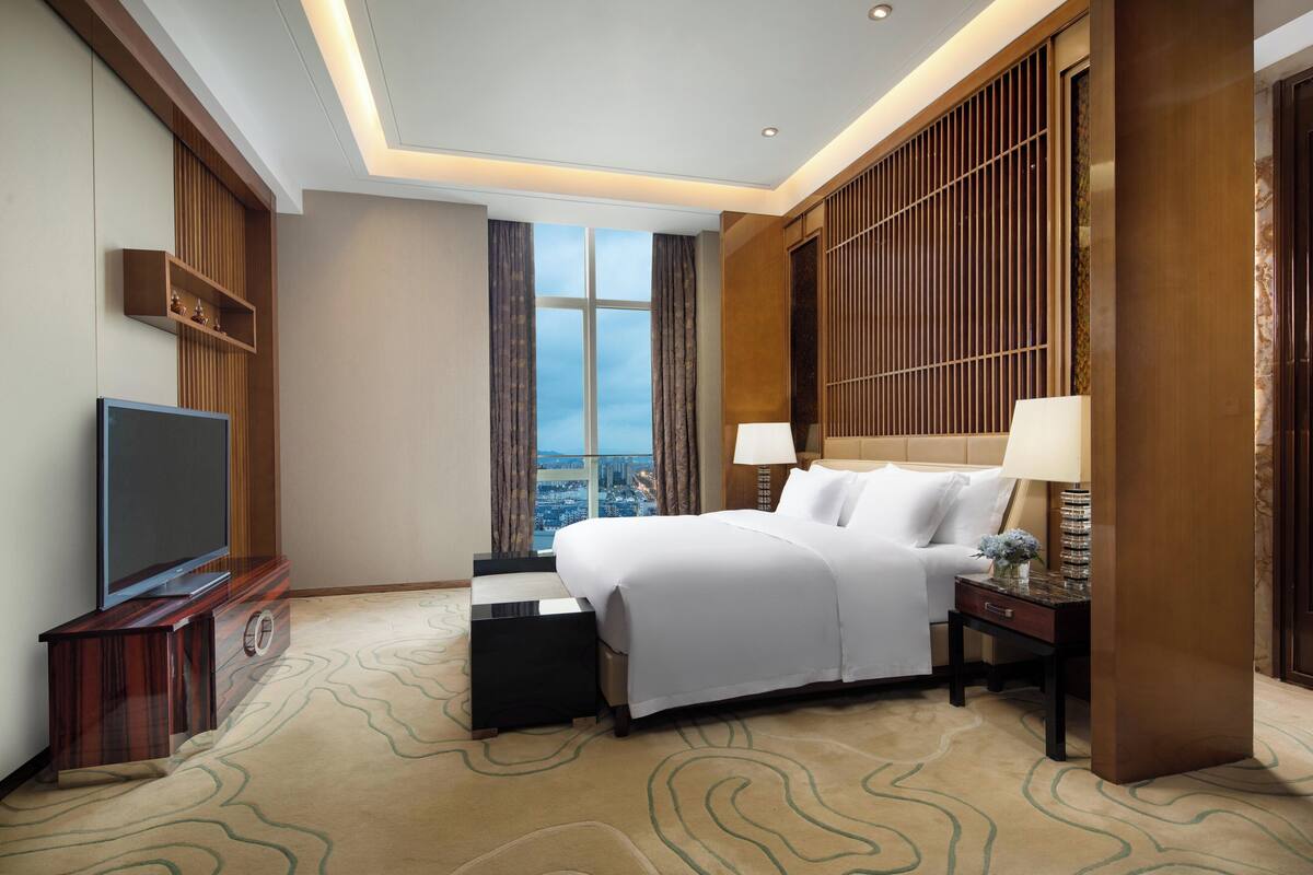 A hotel room with a large bed, a TV, a nightstand, and a view of the city through the window.