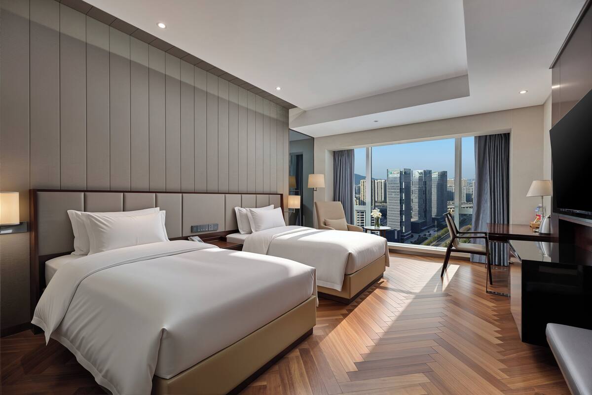 A hotel room with two beds, a TV, a desk with a chair, and a large window with a city view.