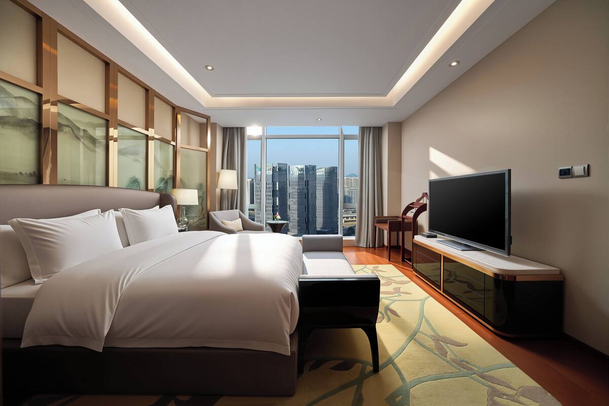 A modern hotel room with a large bed, a flat-screen TV, a seating area, and a view of the cityscape.